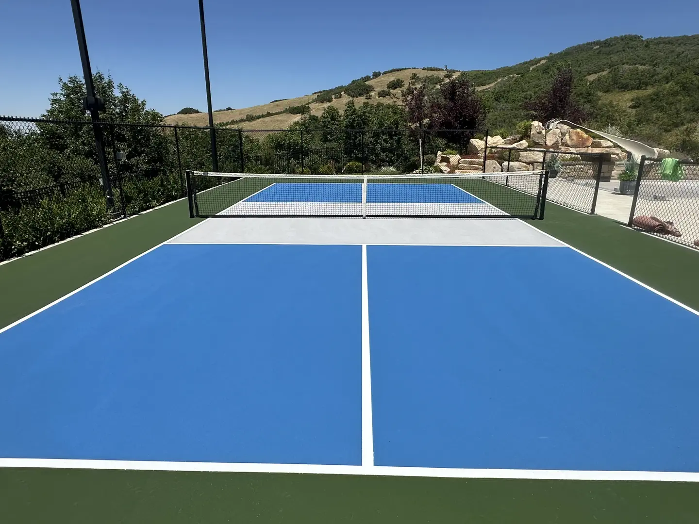 Bright blue pickleball court with green surround and mountain backdrop.