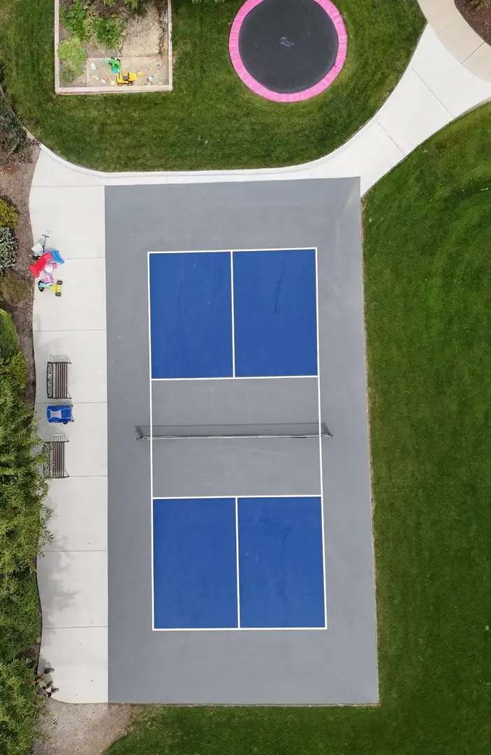Aerial view of a blue pickleball court with gray surroundings and a trampoline nearby.