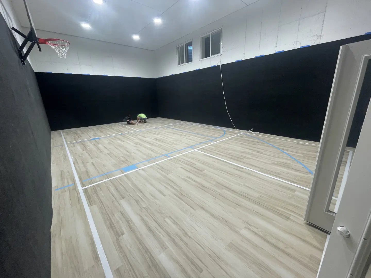 Indoor basketball court with wooden floor and black walls, two people on the floor.