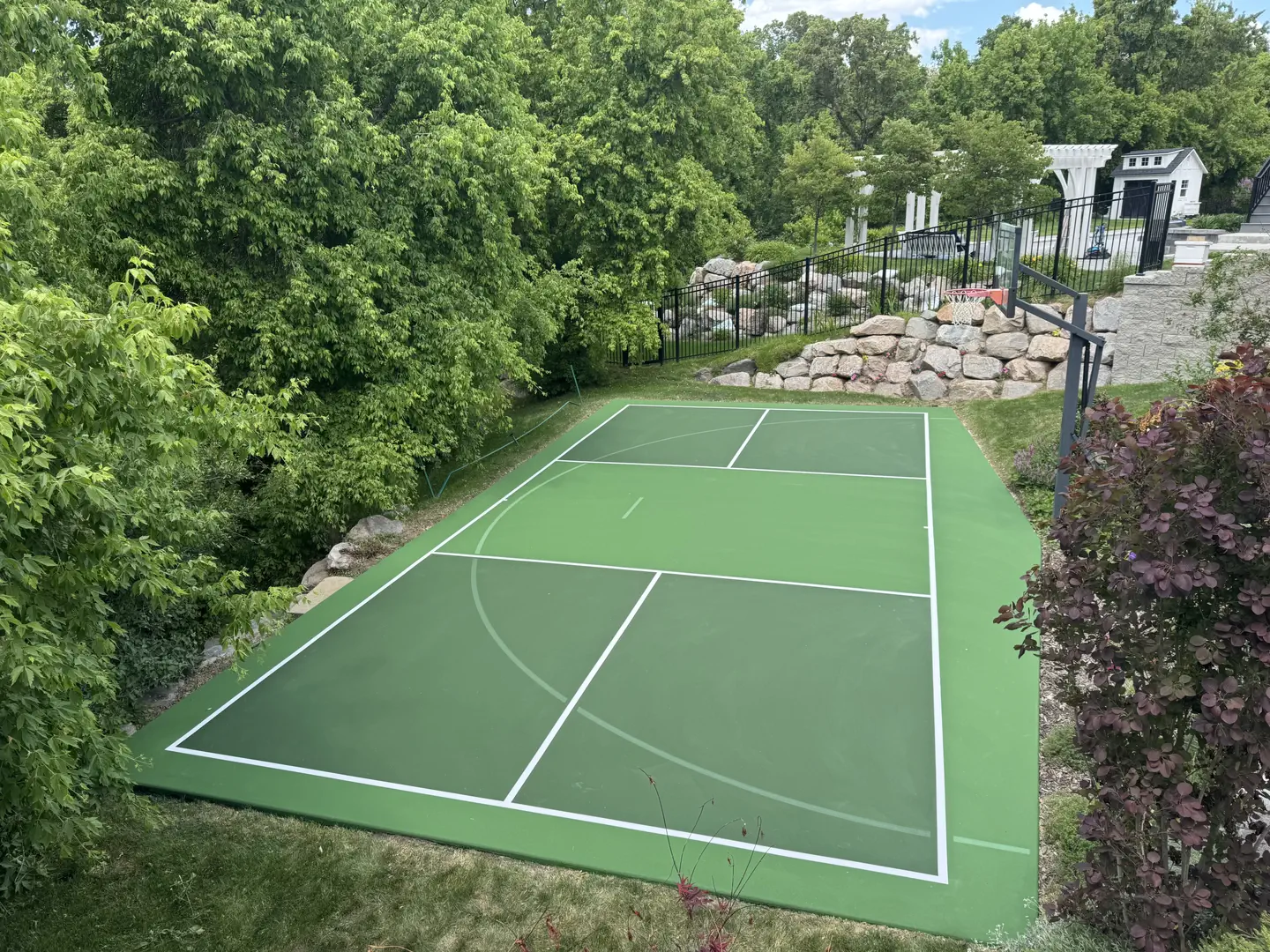Green multi-sport court with basketball hoop in a backyard setting.