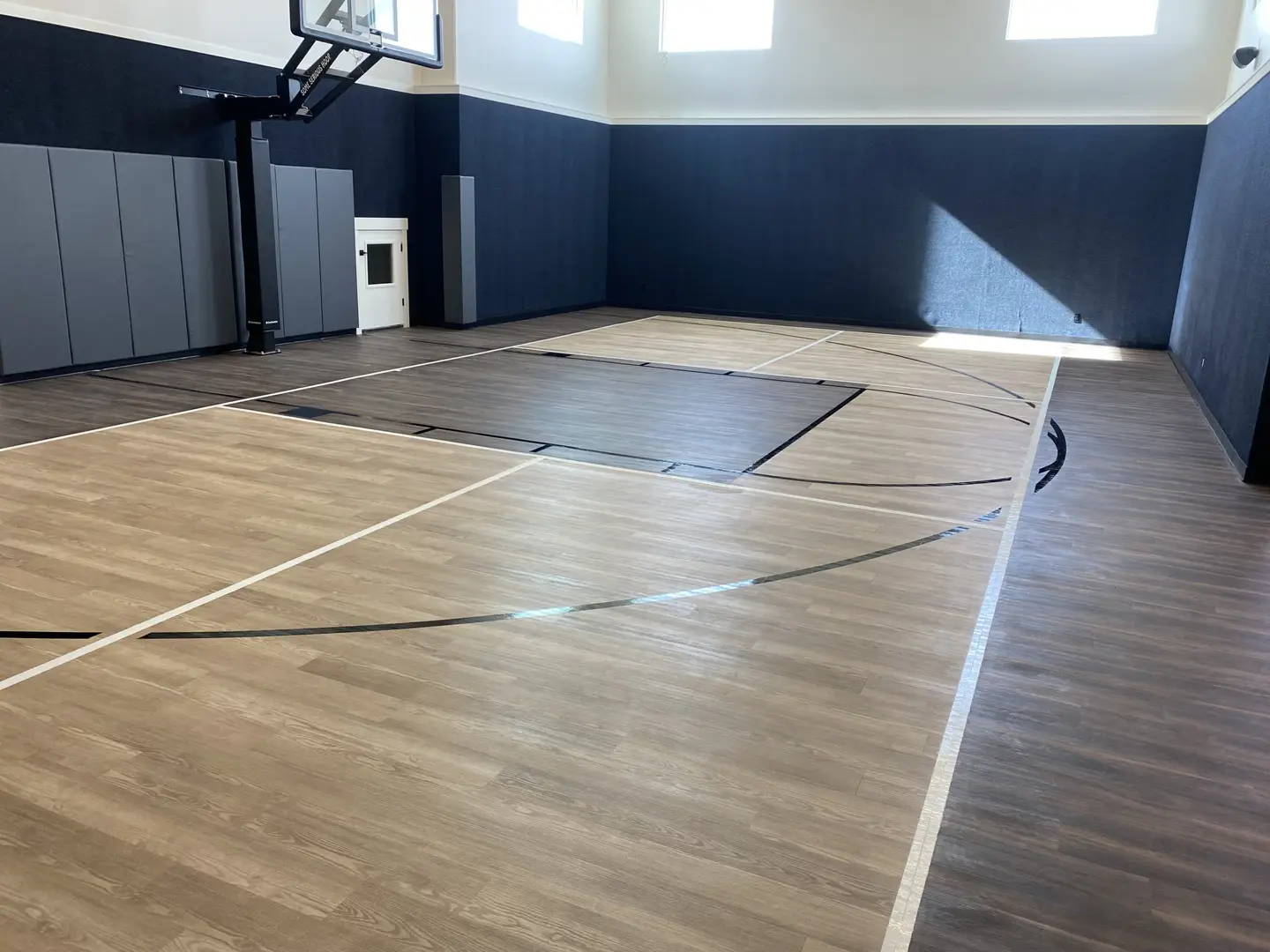 Modern indoor basketball court with wood-look flooring and dark blue walls.