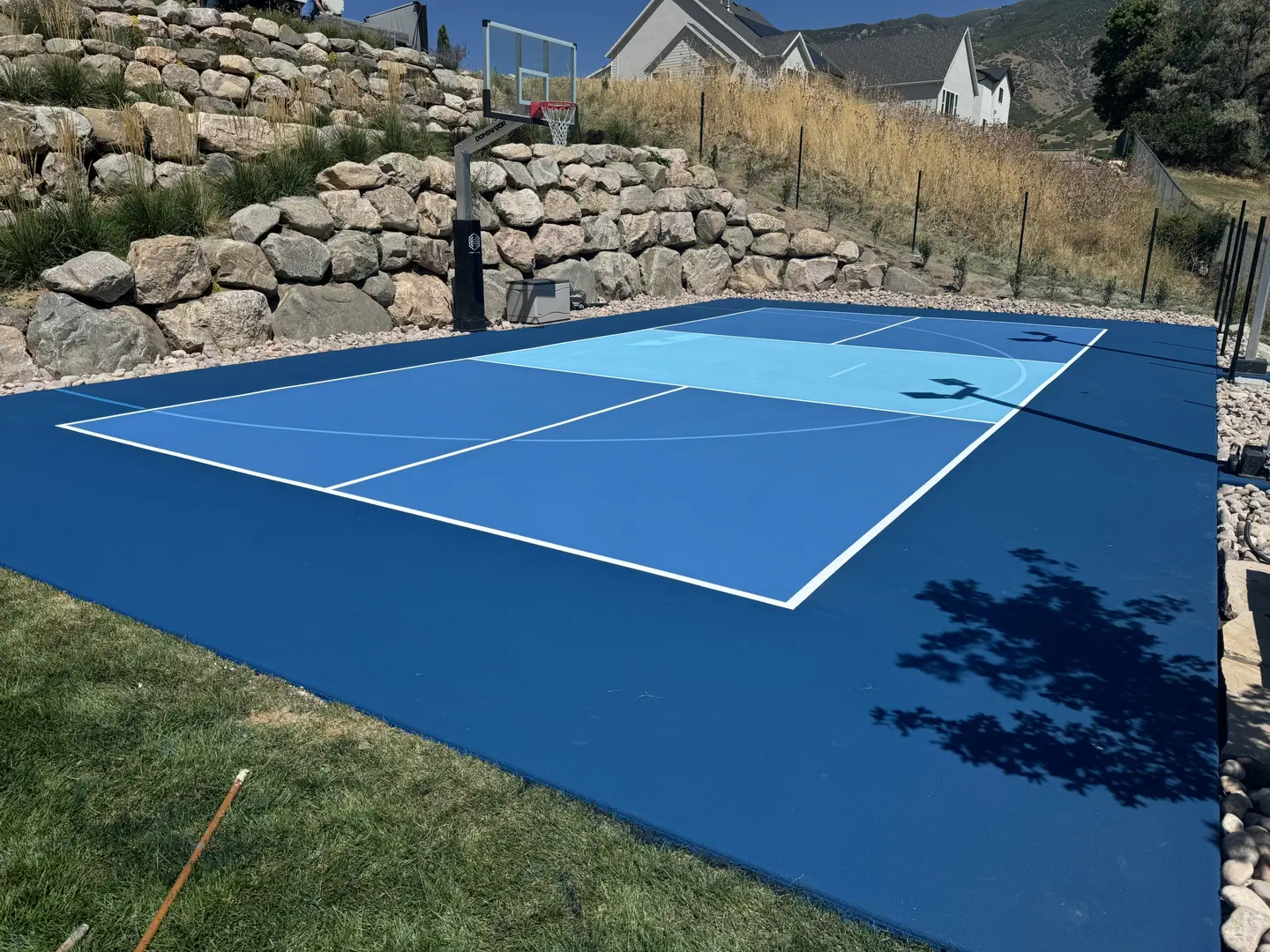 Blue backyard basketball court with a rock wall backdrop.