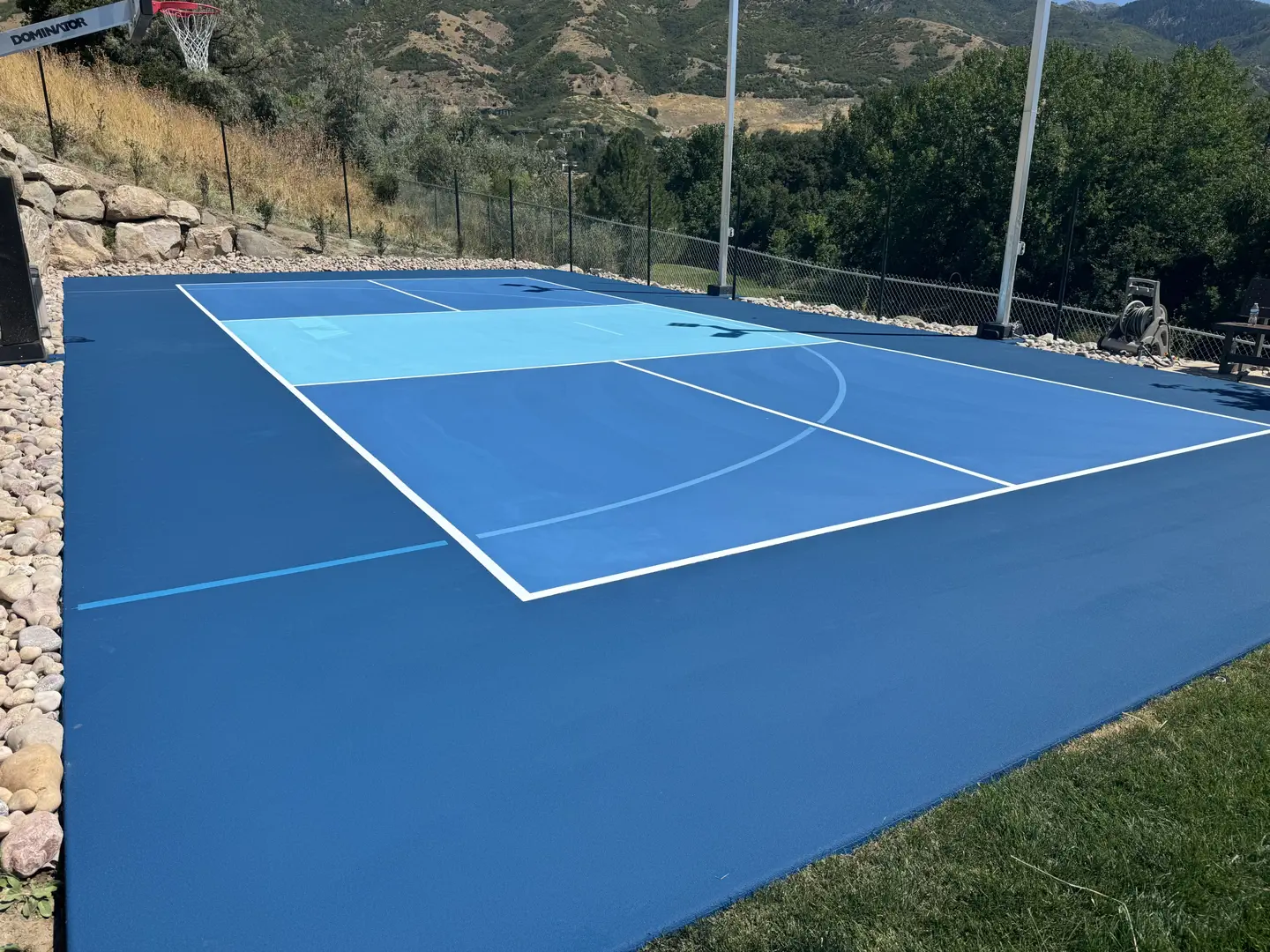 Blue multi-sport court with basketball hoop in a scenic backyard.