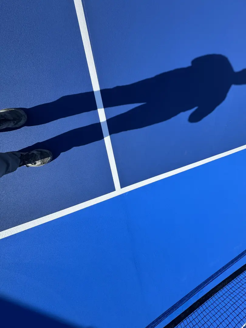 Shadow of a person on a bright blue tennis court with white lines.
