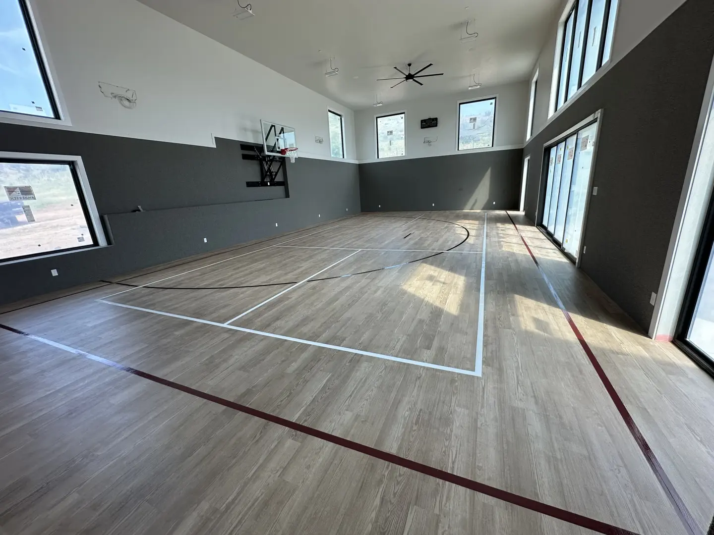 Indoor basketball court with wood-look flooring and large windows.