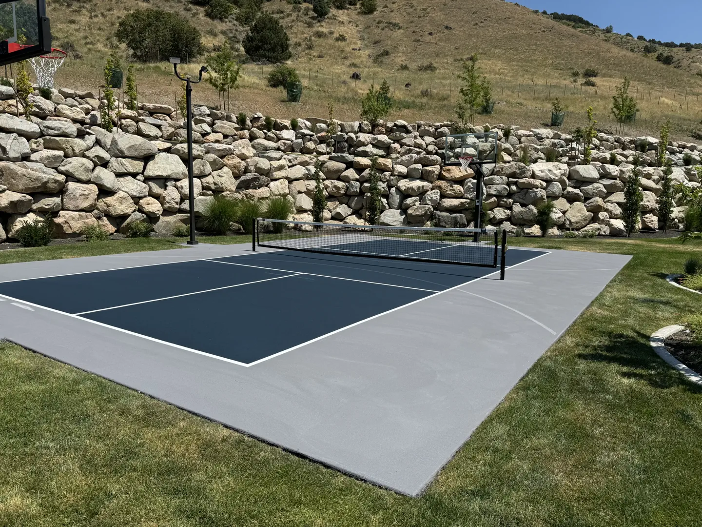 Outdoor pickleball court with basketball hoop and rocky hillside backdrop.