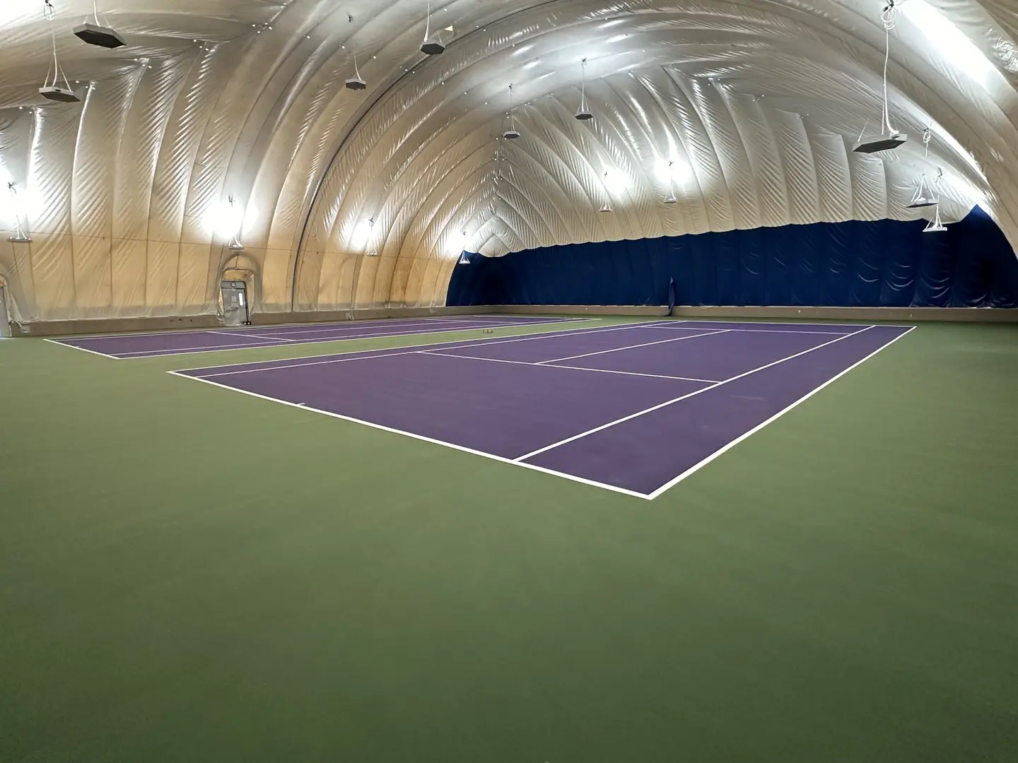 Indoor tennis courts with purple playing surface and green surrounds under an inflatable dome.