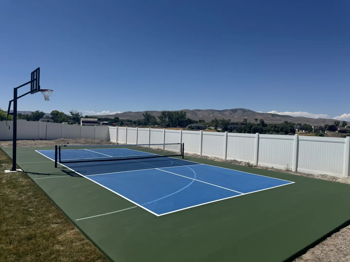 Blue pickleball court with basketball hoop and white fence in a backyard