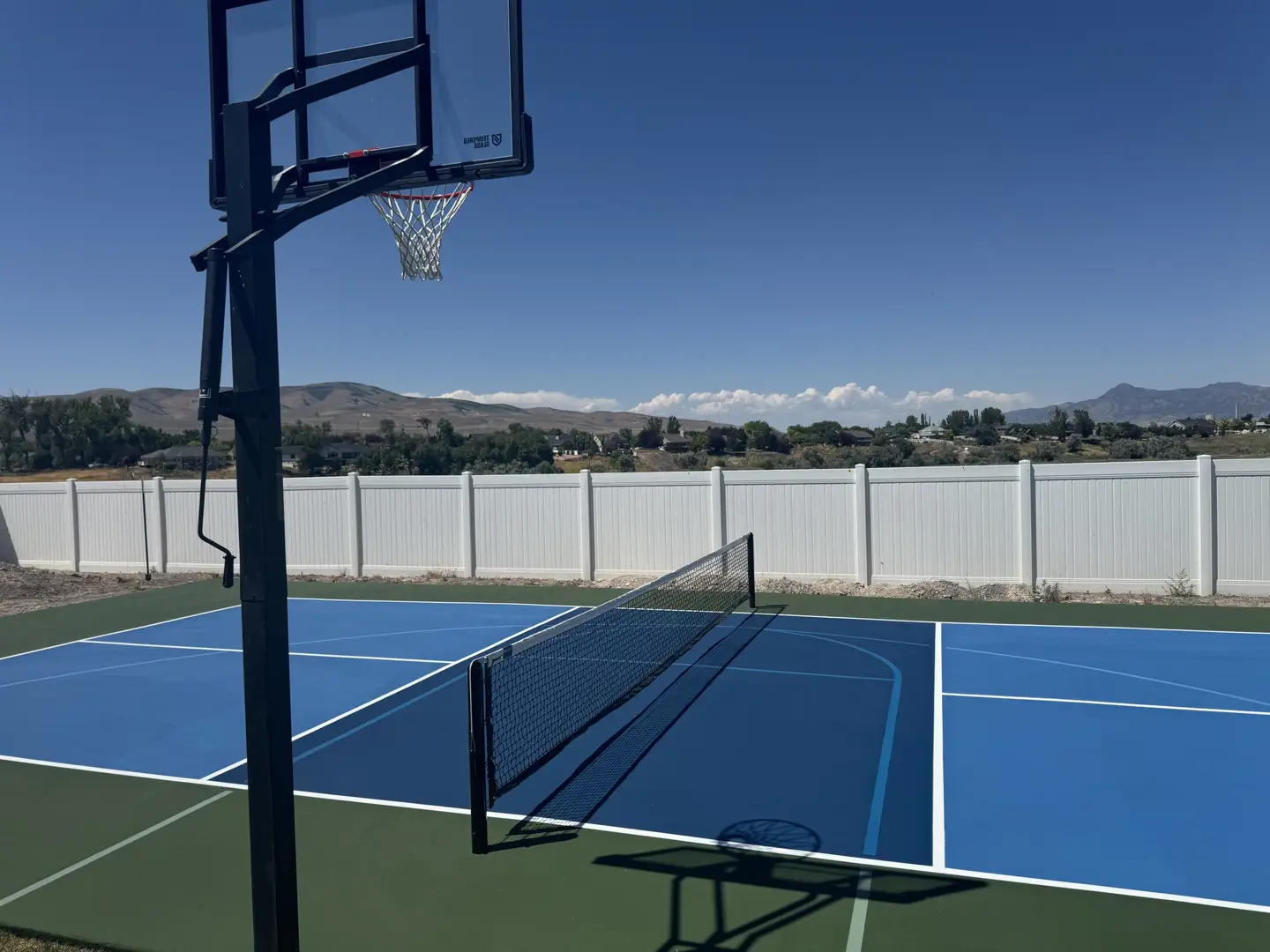 Multi-sport court with basketball hoop and net, blue and green surface, mountain backdrop.