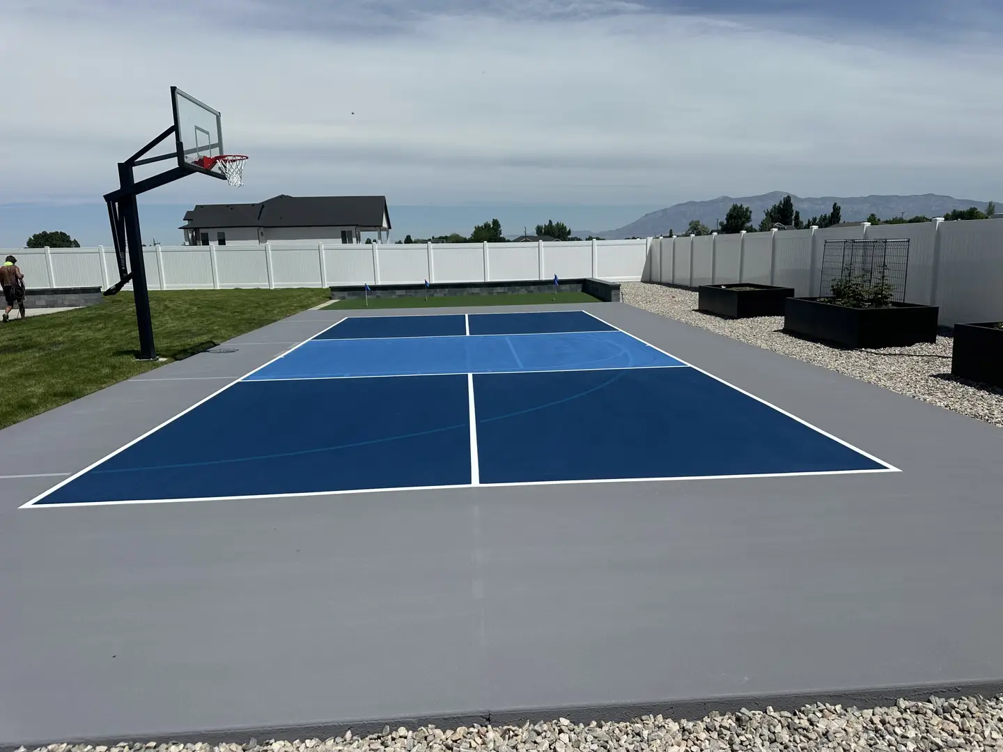 Newly painted blue and gray pickleball court with basketball hoop in backyard.